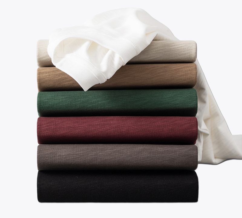 a neatly stacked collection of crew neck t-shirts in different colors, including white, red, green, brown, and black, showcasing the fabric's texture and variety.