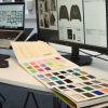 A designer’s workstation featuring a computer monitor displaying digital fashion design sketches, with a large, open color-matching swatch book spread across the desk to facilitate precise material and color selection during the apparel product development process.