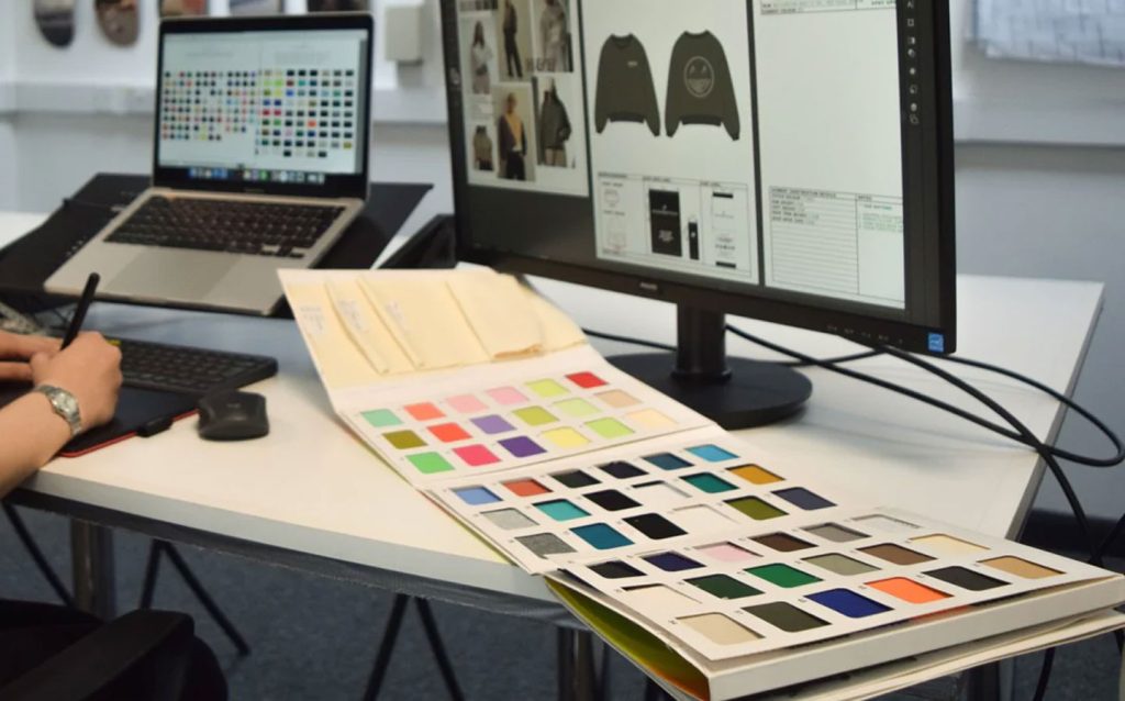 A designer’s workstation featuring a computer monitor displaying digital fashion design sketches, with a large, open color-matching swatch book spread across the desk to facilitate precise material and color selection during the apparel product development process.