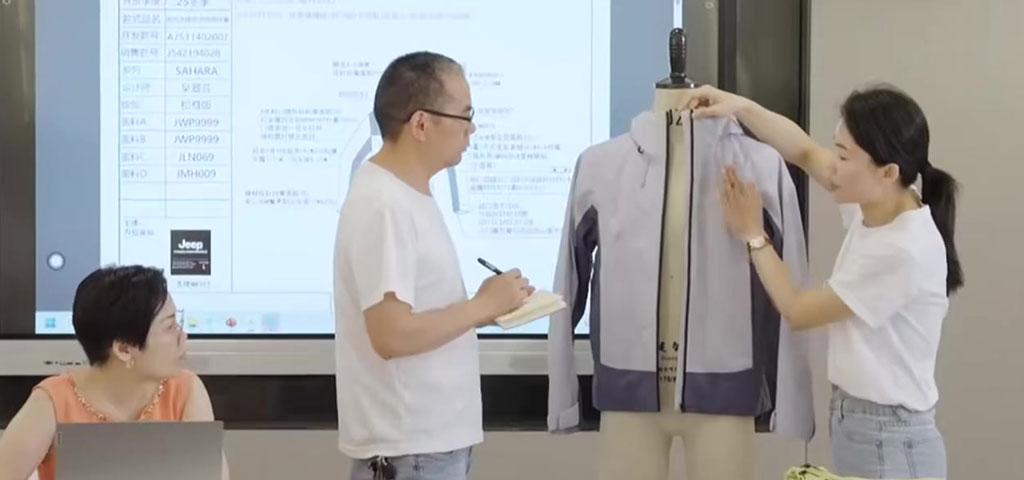 A team of professionals conducting a garment prototype fitting session in a meeting room, where they examine a light grey jacket on a mannequin. The team members are reviewing technical specifications on a large display screen behind them, ensuring the fit, design, and construction details align with the development standards.