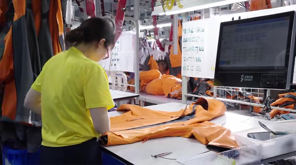 A garment quality control inspection process in a manufacturing facility, featuring a worker in a bright yellow uniform meticulously checking the seams and zippers of an orange waterproof jacket on a workstation. A computer screen displays real-time production data, highlighting the company’s commitment to quality assurance and detailed garment examination before final approval.