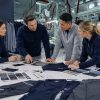 A collaborative meeting between a diverse professional apparel sourcing team and manufacturers at a production facility, reviewing technical garment specifications, fabric swatches, and design sketches on a large table to discuss low MOQ production requirements.