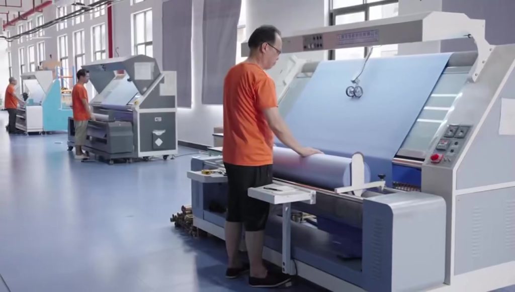A technician performing professional fabric inspection in an apparel factory. The worker operates a sophisticated inspection machine to scan rolls of fabric, ensuring material quality, color consistency, and structural integrity before the cutting stage of the garment manufacturing process.