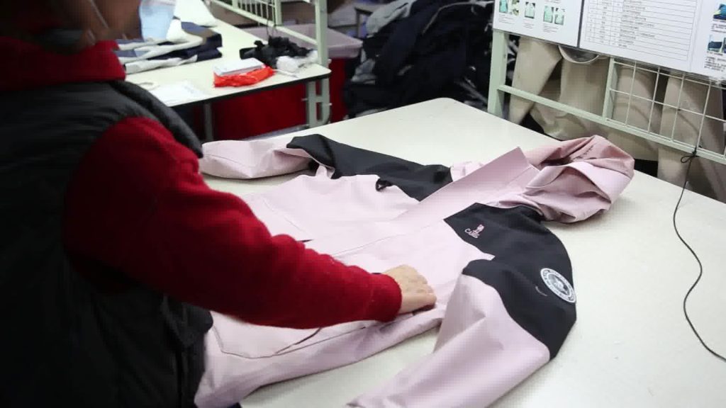 A garment factory worker performing a professional quality control inspection on a pink and black technical jacket, ensuring construction standards are met during the bulk production phase.