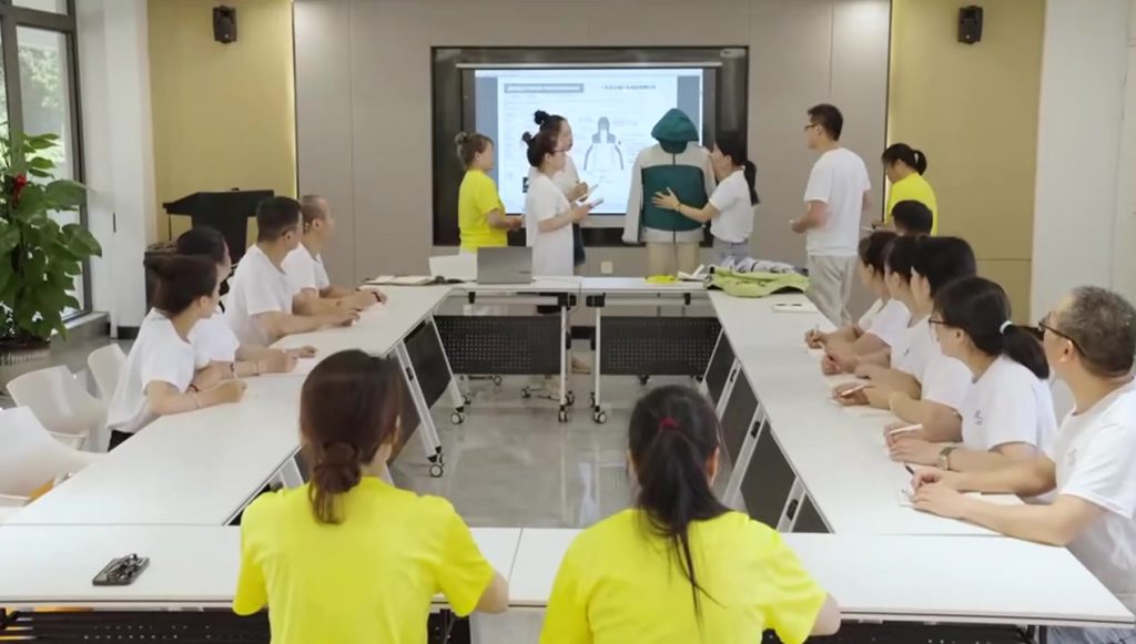 A professional meeting room environment where a team is gathered around a table to conduct a Pre-Production Sample (PPS) approval, with members reviewing a garment prototype on a mannequin against a digital presentation.