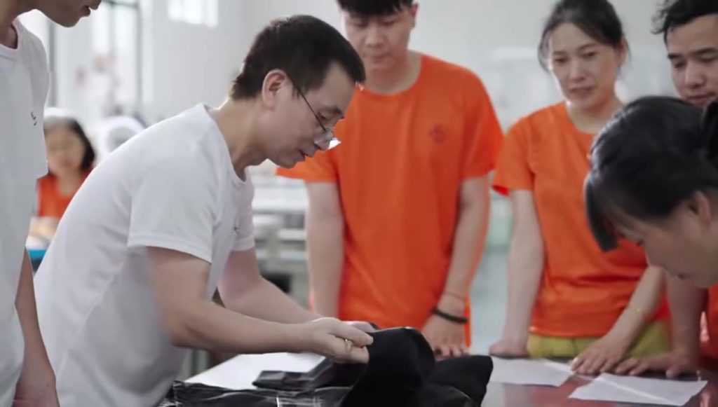 A collaborative team review session in a garment factory, where experienced staff are closely examining and discussing the quality and construction of a black prototype sample garment.