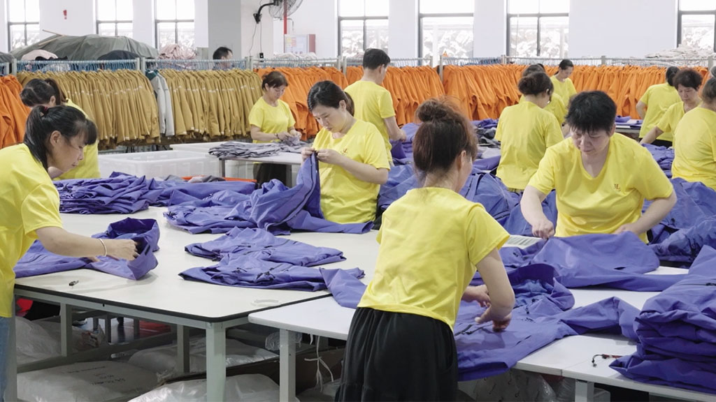 A team of workers in a garment factory performing a rigorous quality control check on a batch of purple jackets, inspecting them on large tables to ensure compliance with production standards before final packaging.