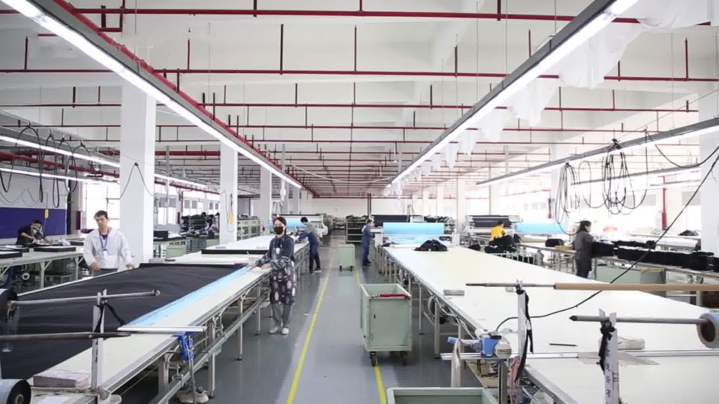 A spacious industrial garment manufacturing facility featuring rows of long cutting tables where skilled workers are actively processing black fabric, demonstrating professional apparel production capabilities.