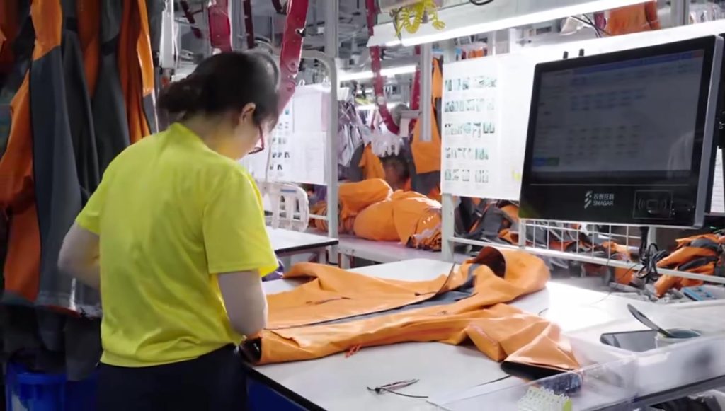 A quality control inspector wearing a yellow shirt carefully checking an orange technical jacket at a workstation equipped with digital monitoring displays in an apparel manufacturing facility.